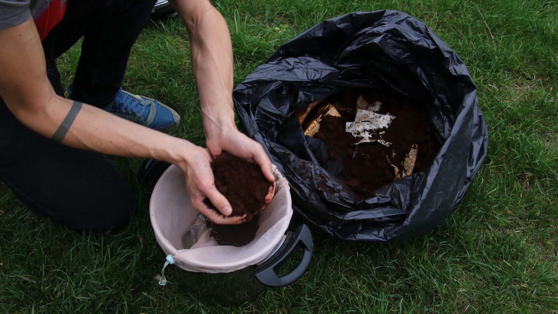 Zwei Hände heben einen Haufen Kaffeesatz aus einem Müllbeutel in einen Pilzeimer.
