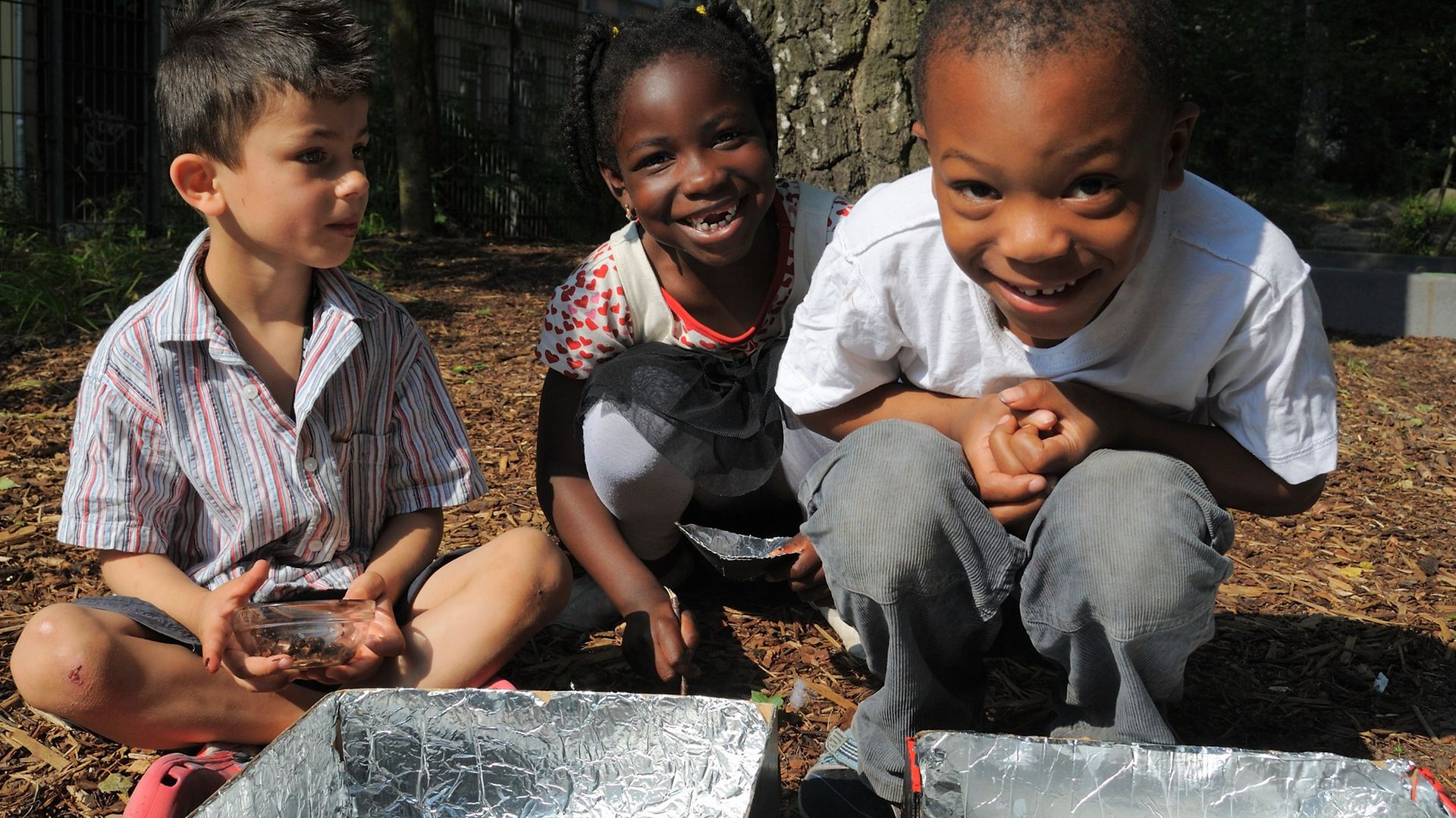Drei Kinder beugen sich lächelnd über selbstgebastelte Solaröfen