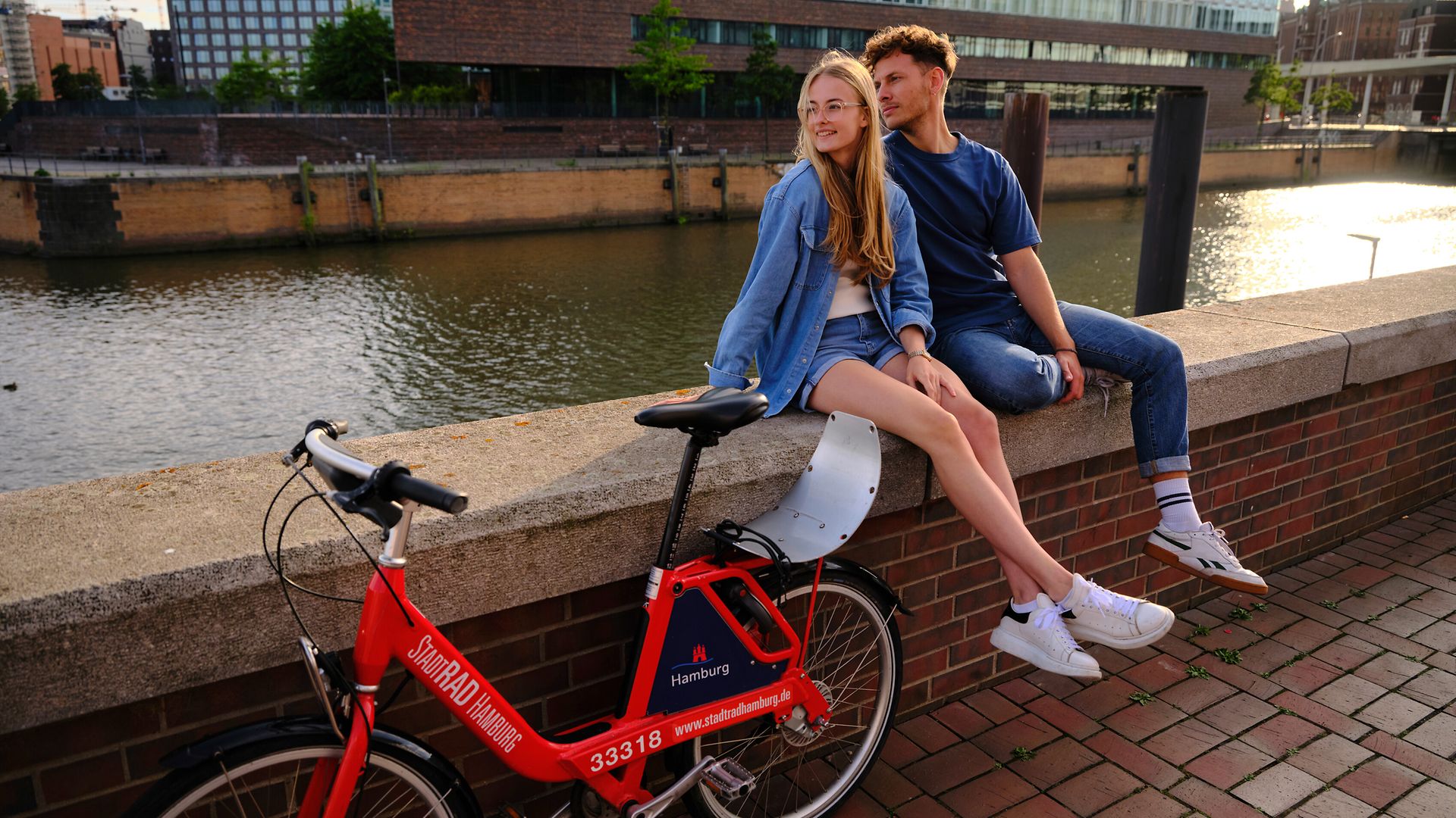 Ein junger Mann und eine junge Frau sitzen auf einer Mauer am Hamburger Oberhafen. Neben ihnen steht ein StadtRAD. 