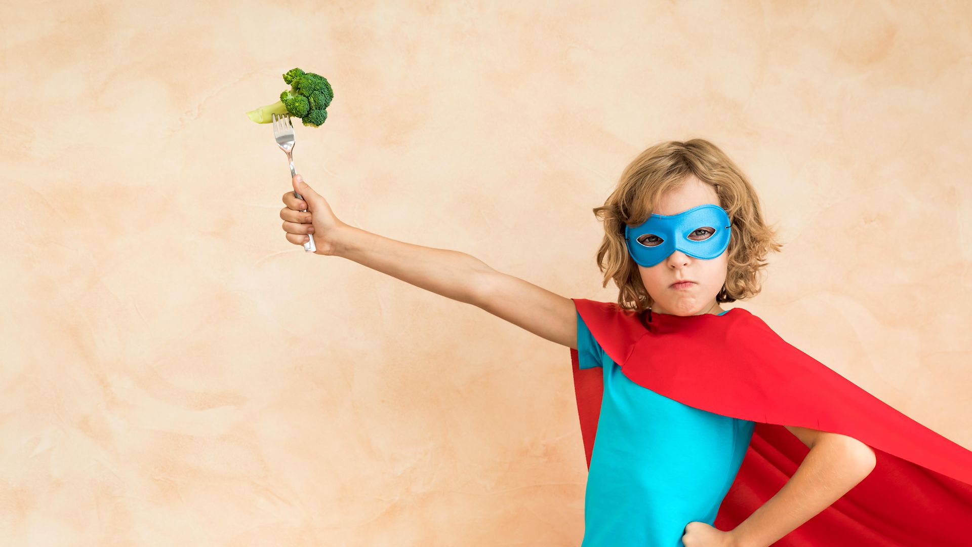Ein Kind mit Maske und Superhelden-Umhang hält eine Gabel hoch, auf der Brokkoli aufgespießt ist.