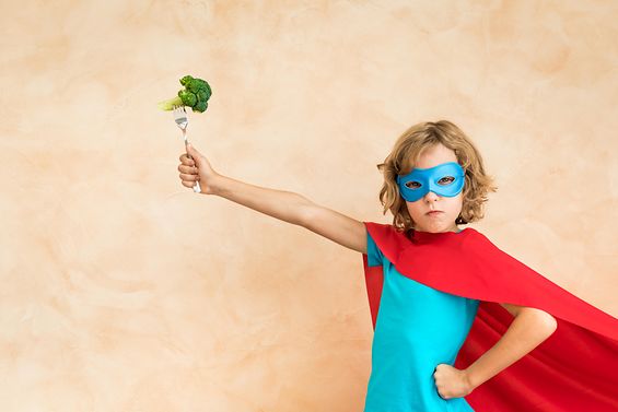 Ein Kind mit Maske und Superhelden-Umhang hält eine Gabel hoch, auf der Brokkoli aufgespießt ist.
