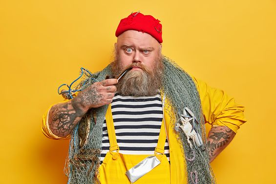 Bärtiger Seemanns mit einer Pfeife im Mund, in die Kamera schauend, posiert mit Fischernetz vor einer gelber Studiowand.