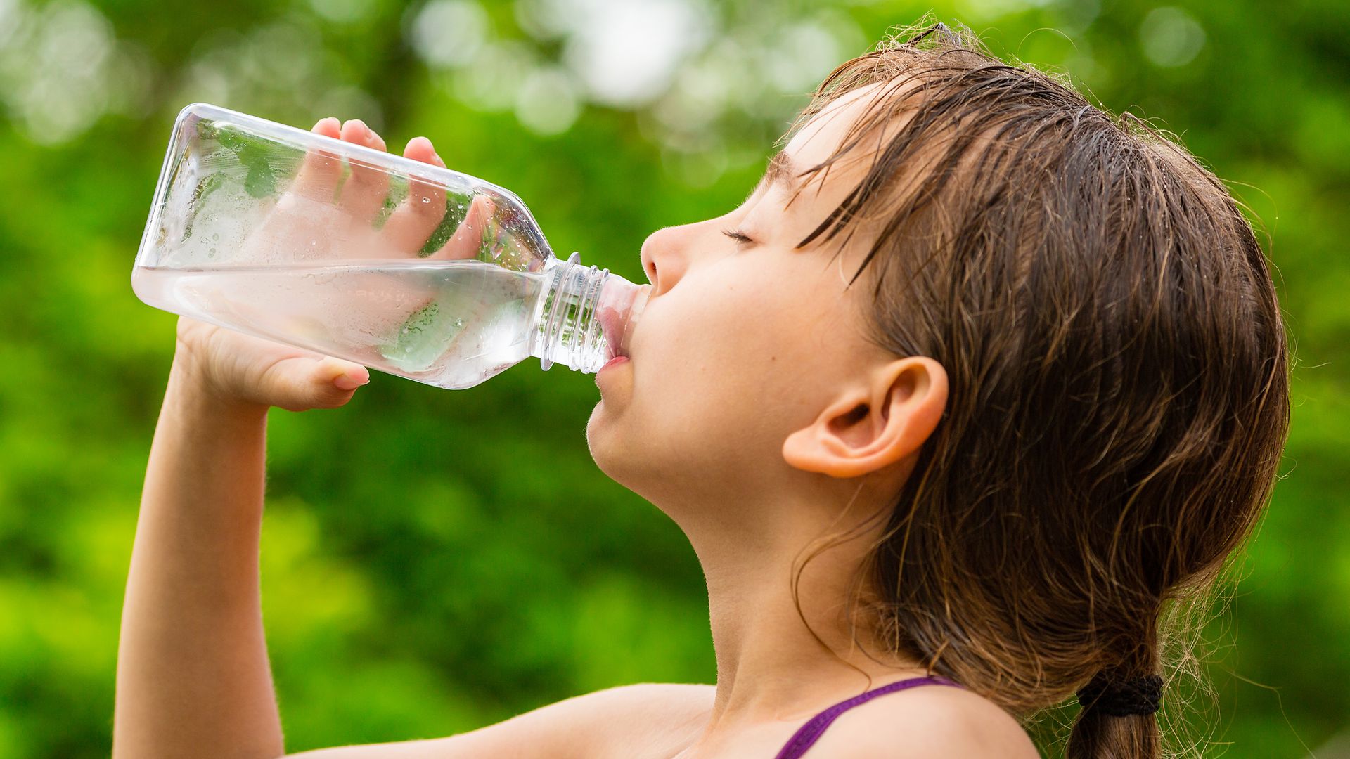Ein Mädchen trinkt mit geschlossenen Augen Wasser aus einer Glasflasche.
