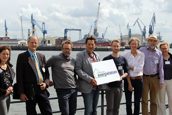 Personen stehen vor der Kulisse das Hamburger Hafens und halten ein Schild mit der Aufschrift „Hamburger Energielotsen“.